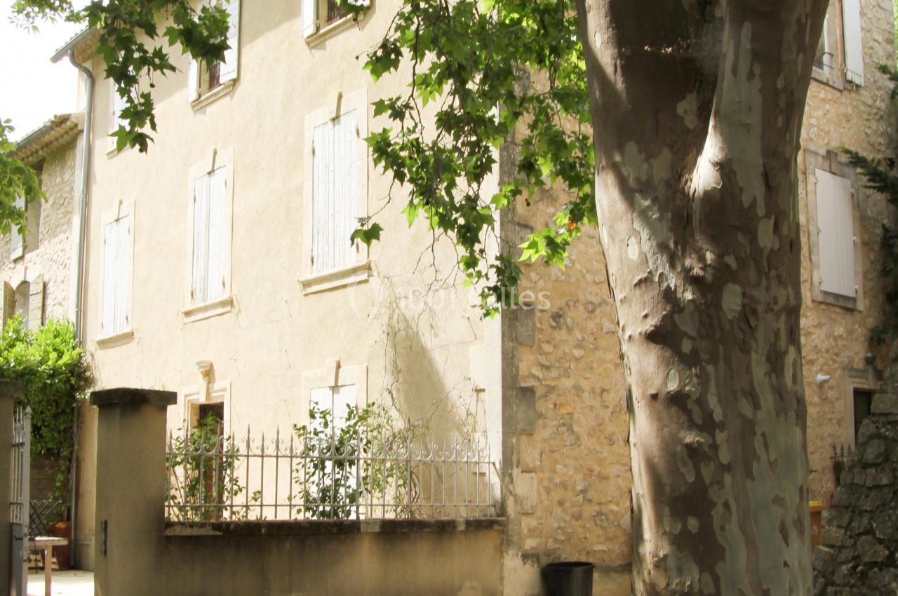 Façade d'une maison en pierre avec volets fermés, partiellement ombragée par un grand platane au premier plan.