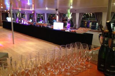 Salle de réception sur un bateau avec des tables dressées, des verres à champagne alignés et une vue sur l'eau en arrière…