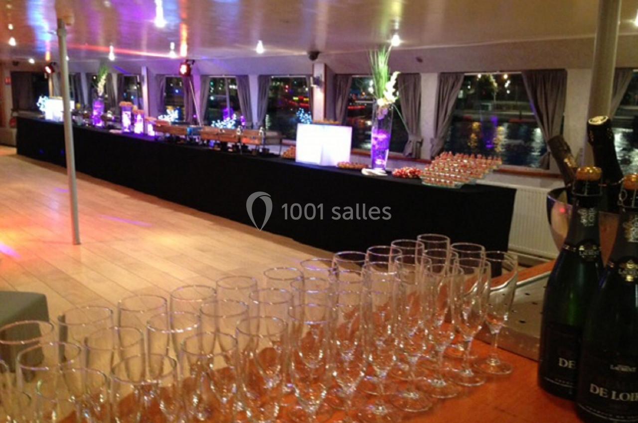 Salle de réception sur un bateau avec des tables dressées, des verres à champagne alignés et une vue sur l'eau en arrière…
