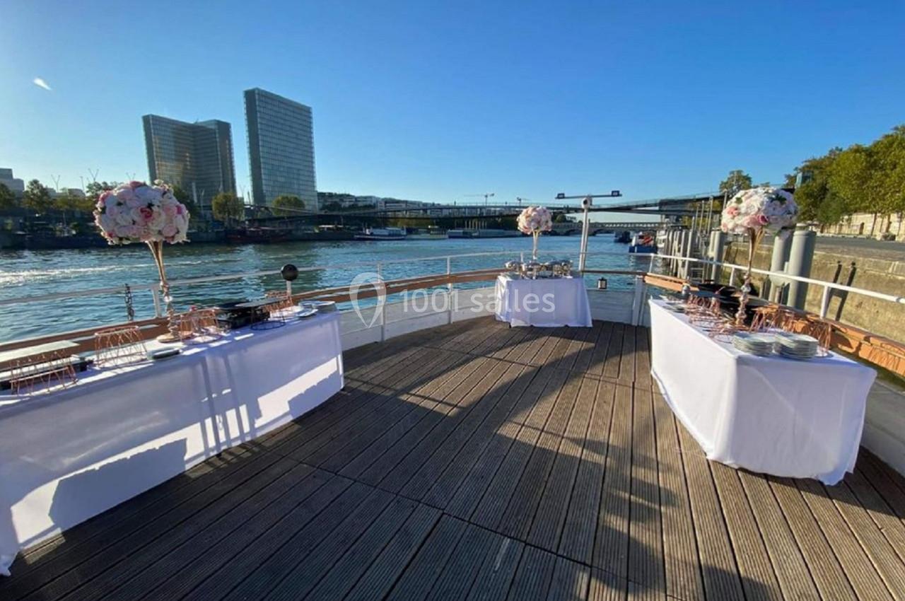 Tables décorées pour un événement sur le pont d'un bateau, avec vue sur la Seine et des immeubles en arrière-plan.