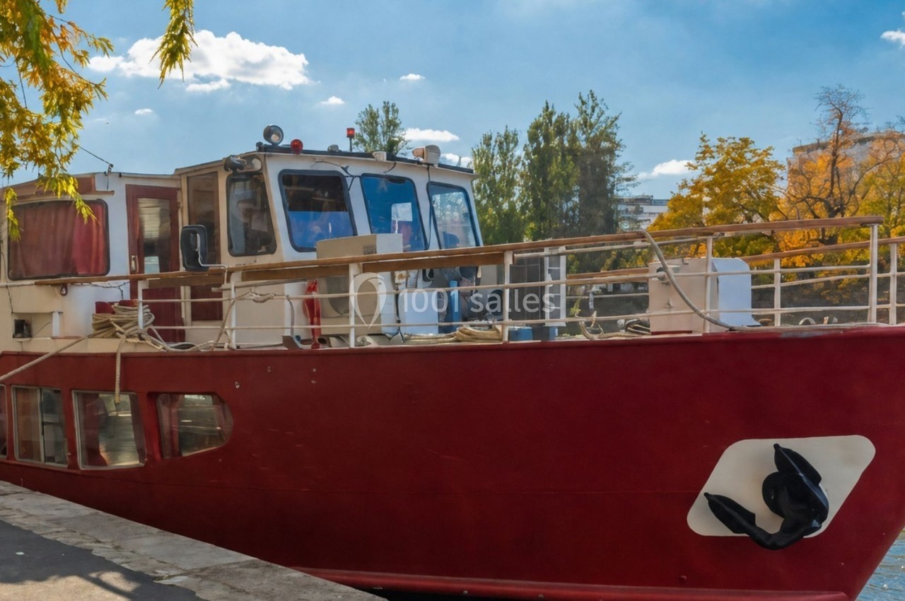 Bateau rouge amarré le long d'une berge, entouré d'arbres aux couleurs automnales et sous un ciel dégagé.