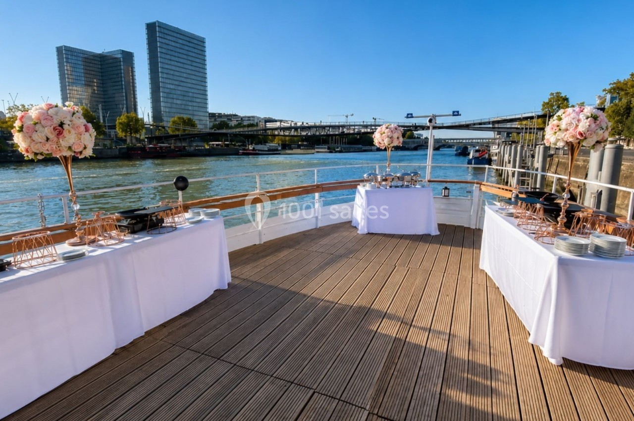 Pont d'un bateau aménagé pour un événement, avec tables décorées et vue sur un fleuve bordé de bâtiments modernes.