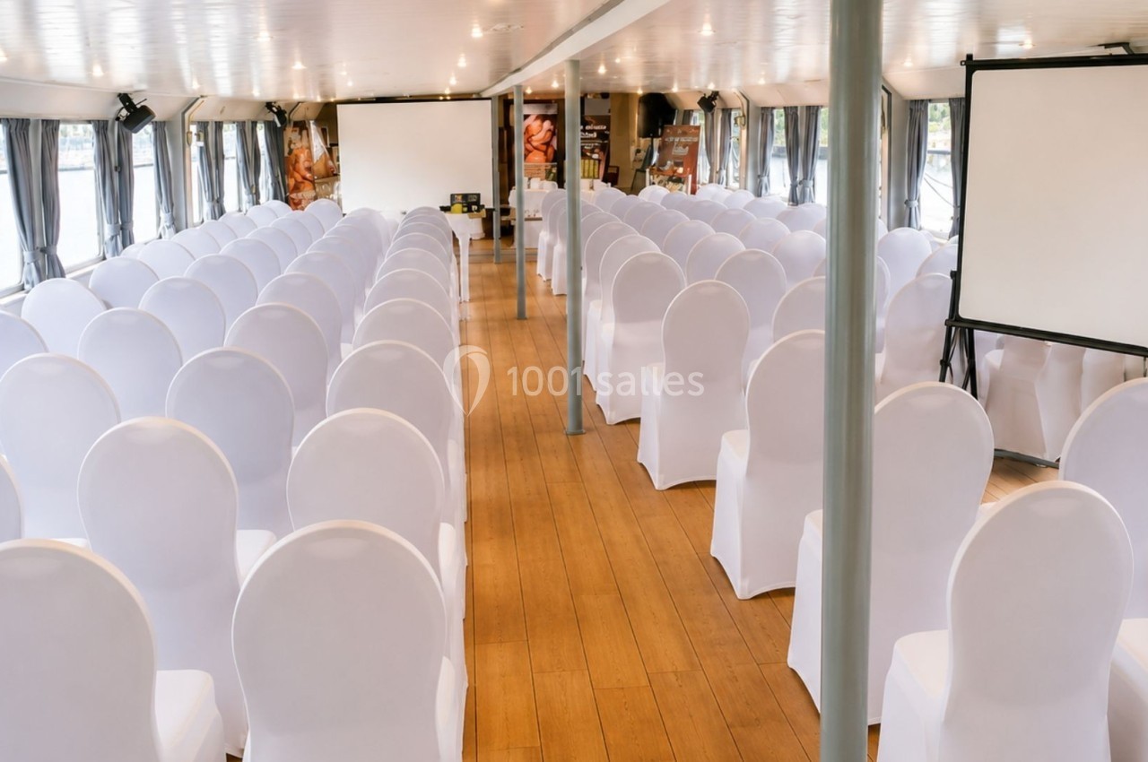 Salle de conférence sur un bateau avec des chaises blanches alignées, un écran de projection et des fenêtres donnant sur l…
