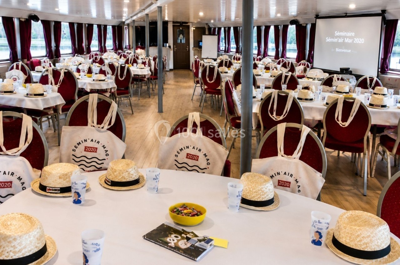 Salle de séminaire sur un bateau, tables dressées avec chapeaux, sacs en toile et accessoires promotionnels.