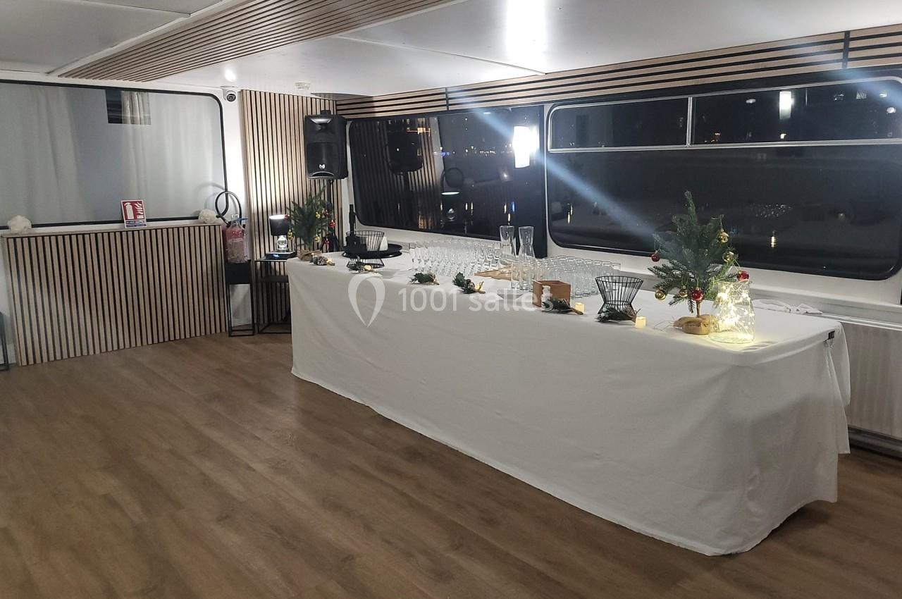 Salle avec parquet, table longue décorée de plantes et lumières, grandes fenêtres donnant sur un extérieur nocturne.