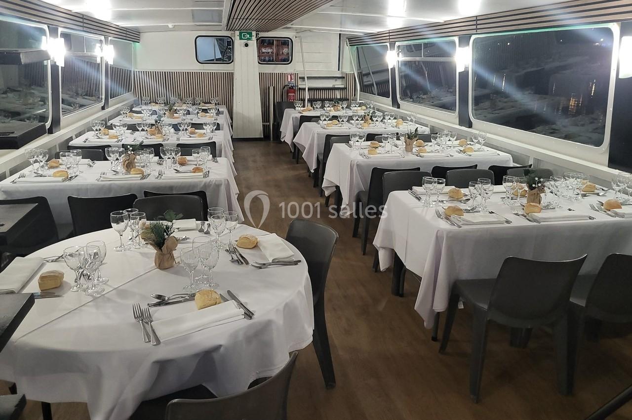 Salle de réception sur un bateau, avec tables dressées pour un dîner, nappes blanches et décoration sobre.