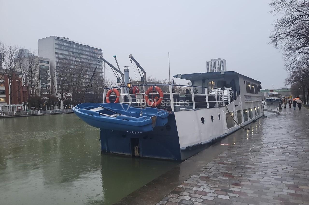 Bateau amarré le long d'un canal par temps gris, avec des immeubles et des arbres en arrière-plan.