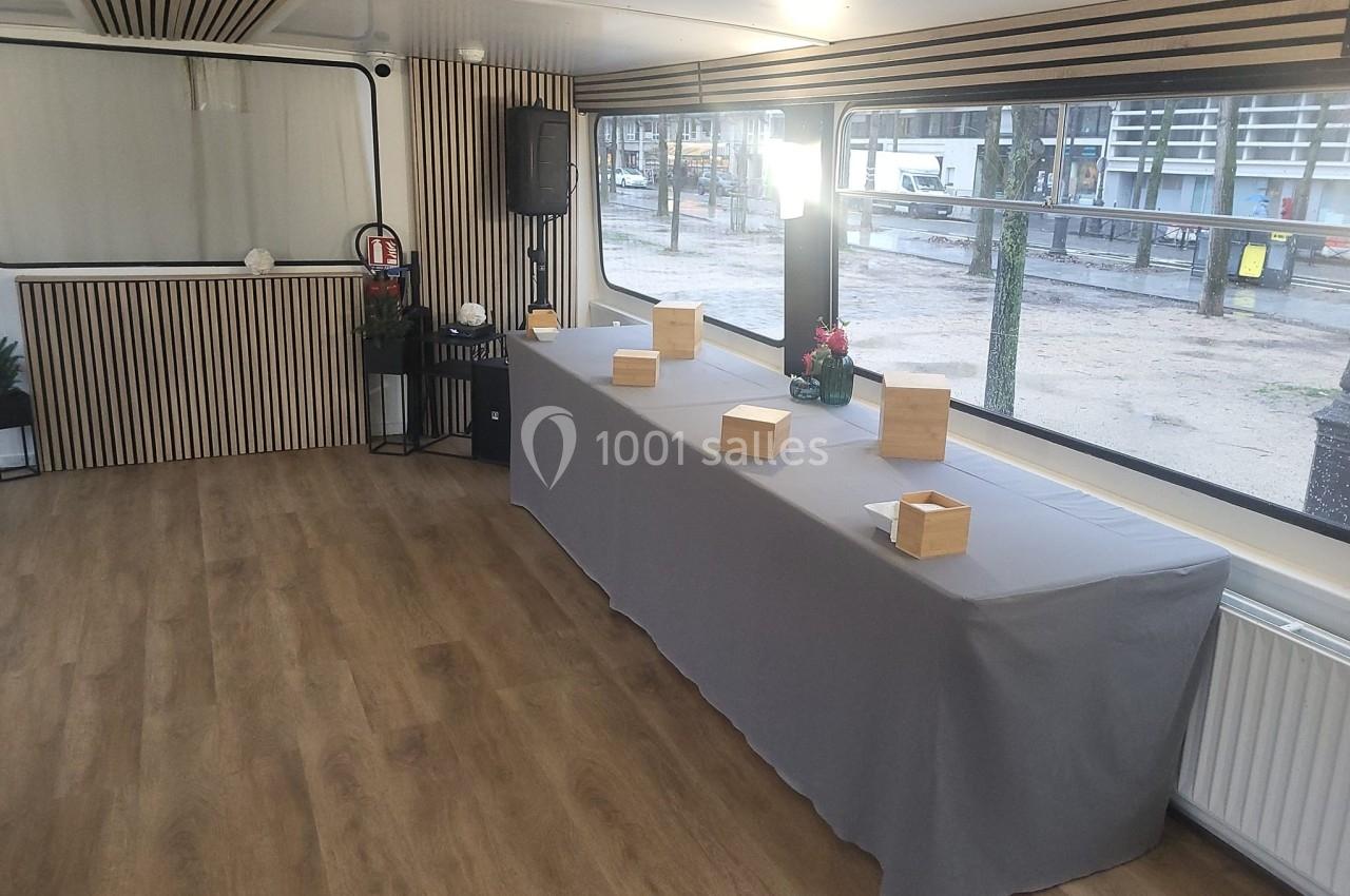Salle lumineuse avec parquet, table recouverte d'une nappe grise, boîtes en bois et plantes en décoration.