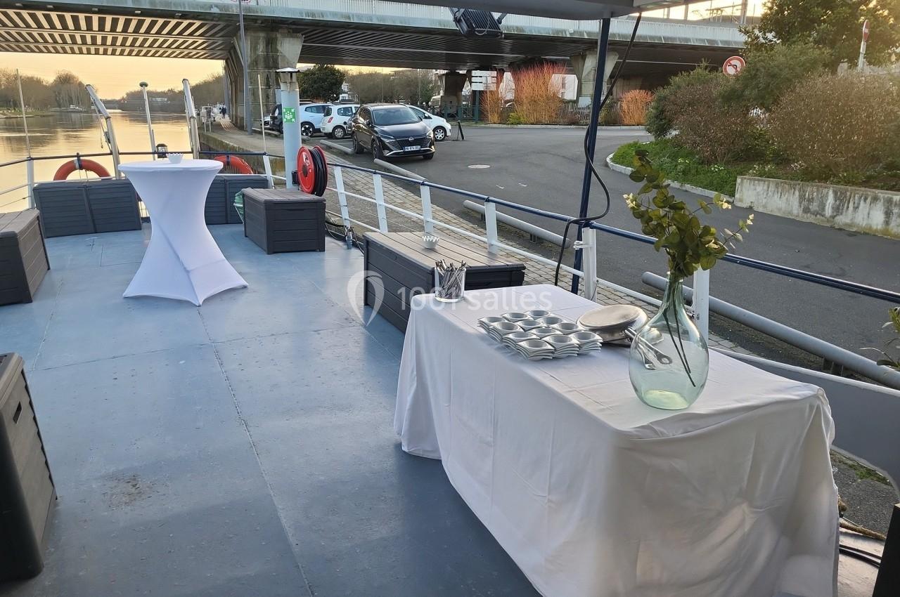 Table dressée avec vaisselle et vase, sur le pont d'un bateau amarré près d'une route et d'un pont.