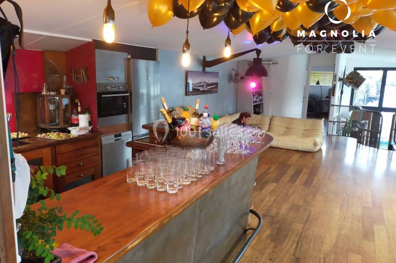 Comptoir en bois avec verres et seau à champagne, décoré de ballons noirs et dorés dans une salle lumineuse.