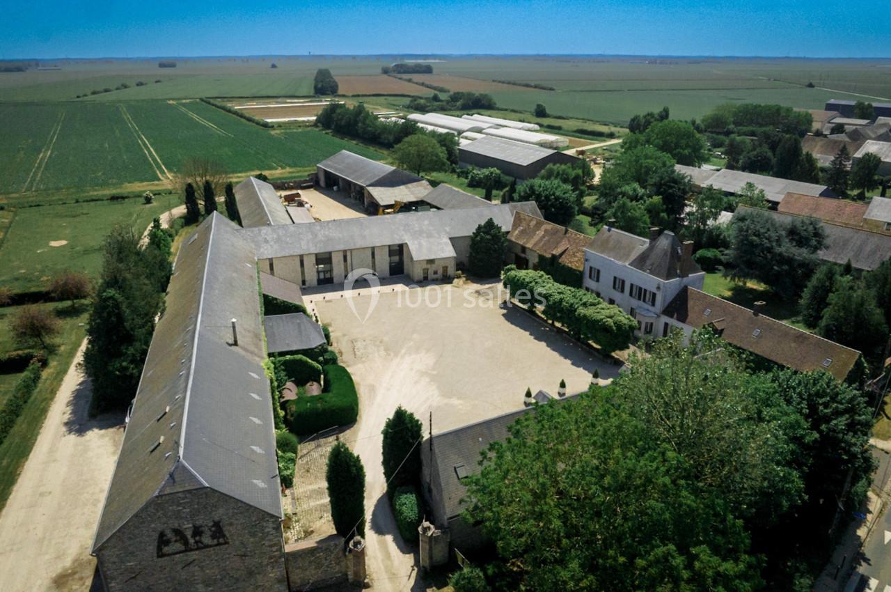 Vue aérienne d'une ferme avec bâtiments en pierre, cour centrale et champs environnants sous un ciel dégagé.