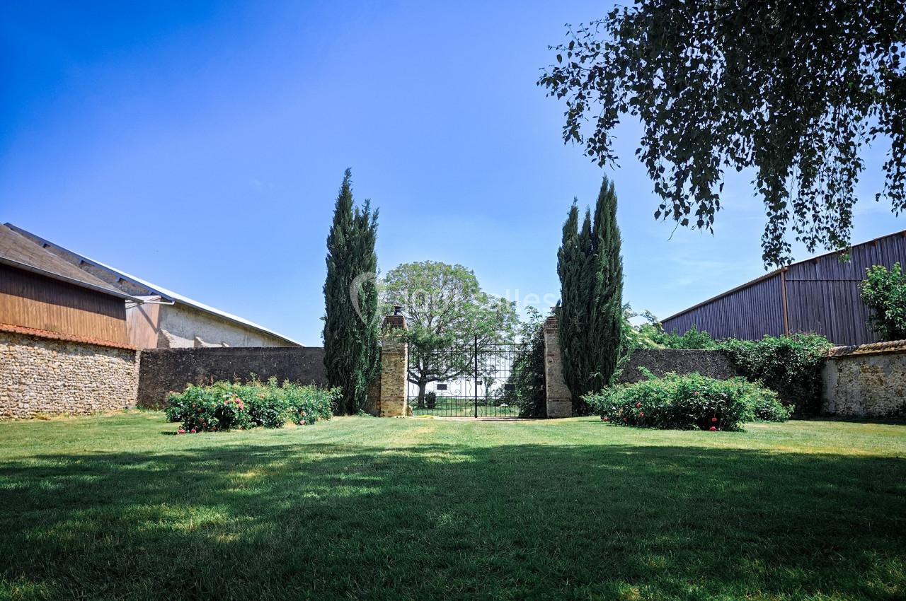 Jardin verdoyant avec pelouse, parterres de fleurs, cyprès et portail en fer forgé sous un ciel bleu dégagé.