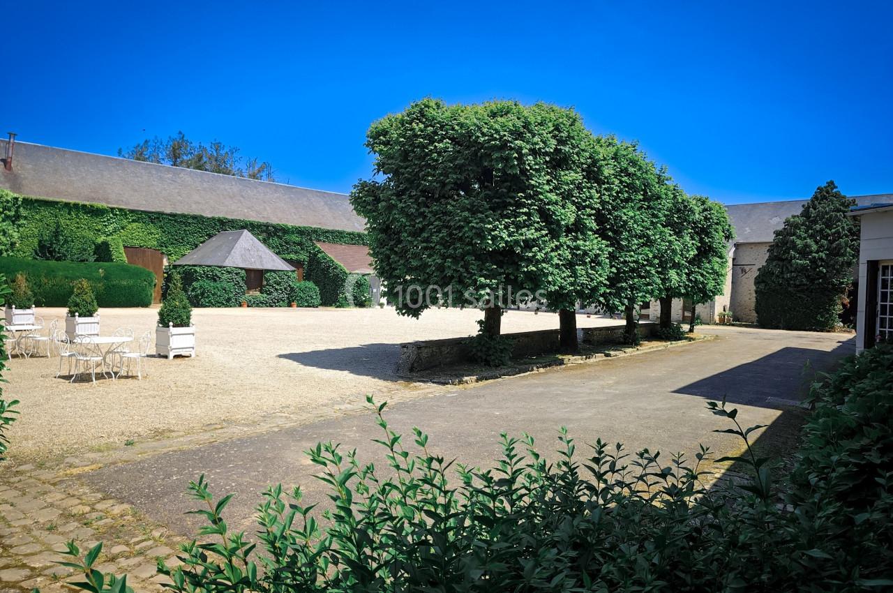 Cour extérieure avec pavés, arbres taillés, chaises blanches et bâtiments en pierre recouverts de végétation.