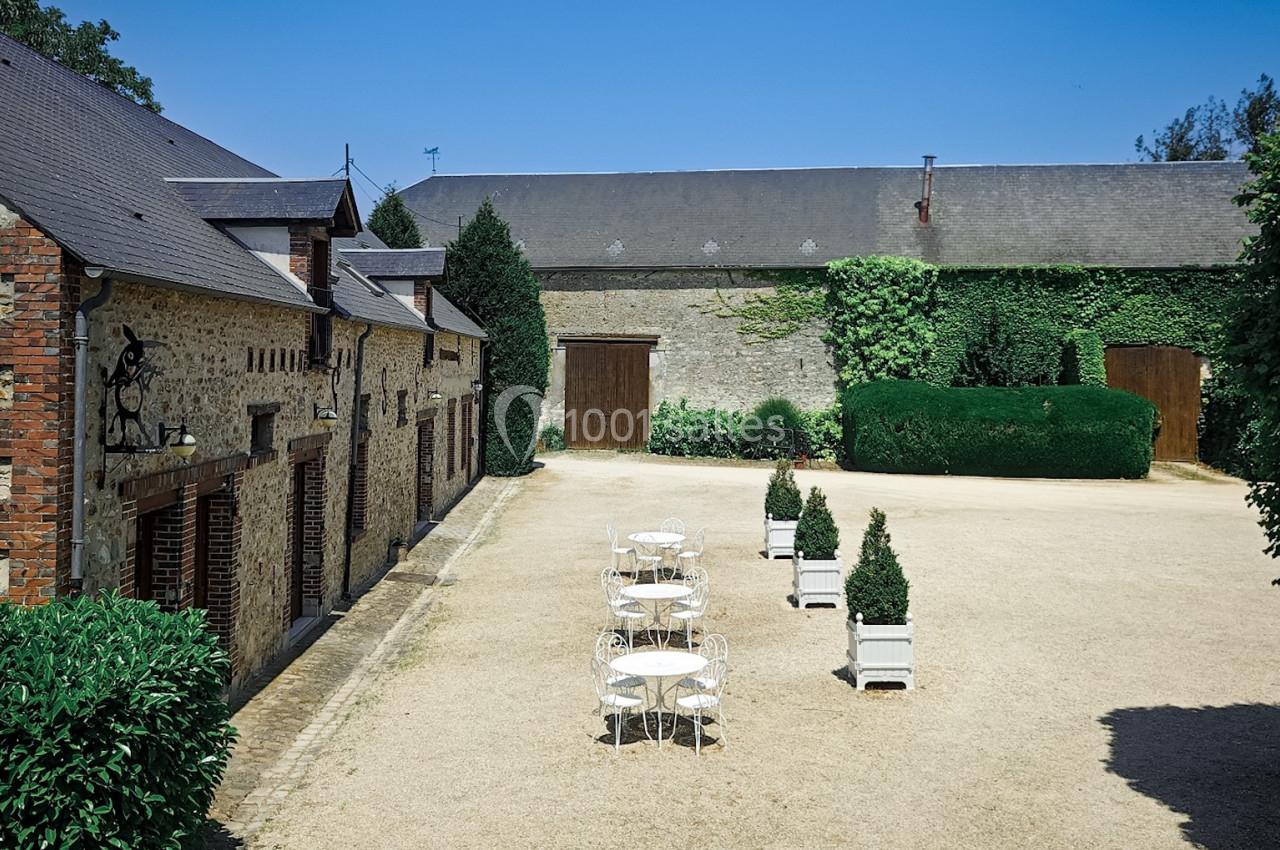 Cour intérieure pavée avec tables et chaises blanches, entourée de bâtiments en pierre et de verdure sous un ciel bleu.