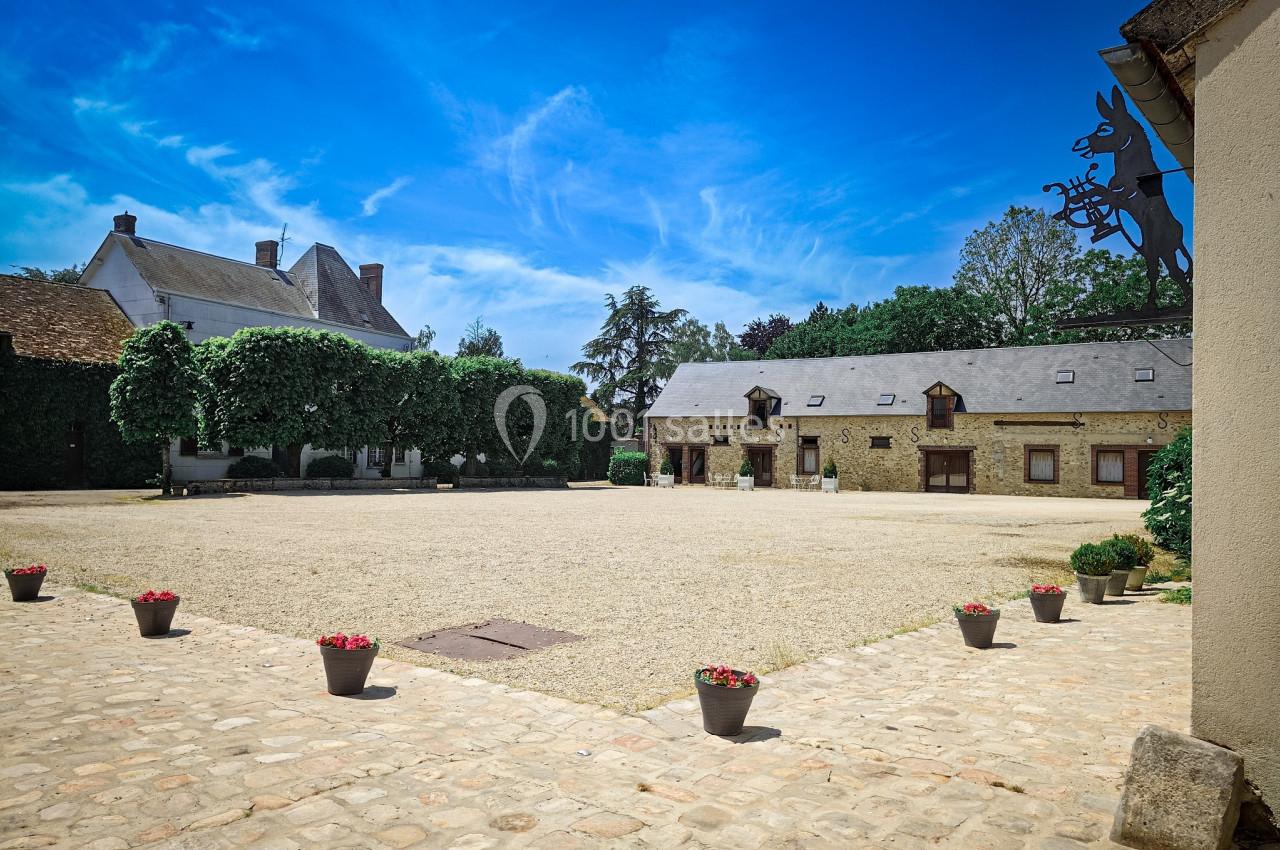 Cour pavée bordée de bâtiments en pierre et de végétation, sous un ciel bleu dégagé.
