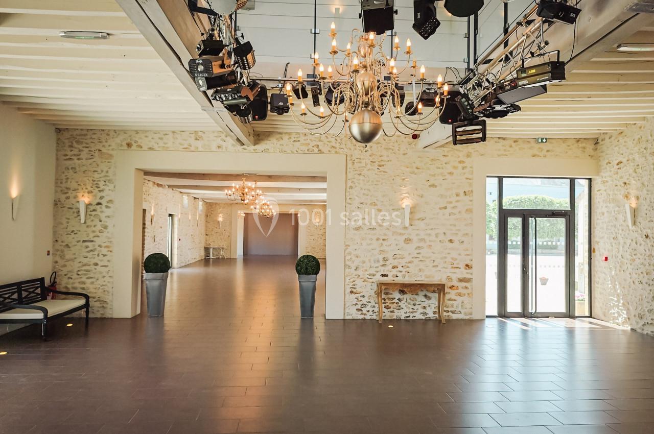 Salle lumineuse avec murs en pierre, lustres suspendus et éclairages, ouverte sur l'extérieur par une porte vitrée.