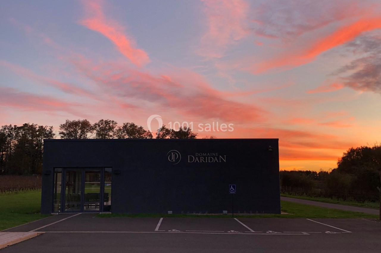 Bâtiment moderne sombre devant un parking vide, au coucher du soleil avec un ciel aux teintes rosées et orangées.