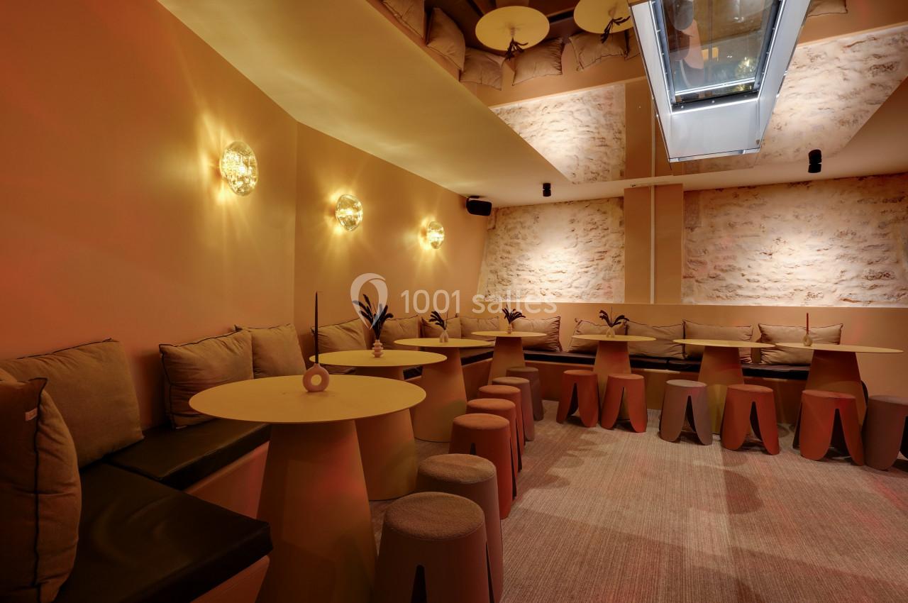 Salle lumineuse avec murs en pierre, banquettes beige, tables rondes et tabourets dans une ambiance chaleureuse.