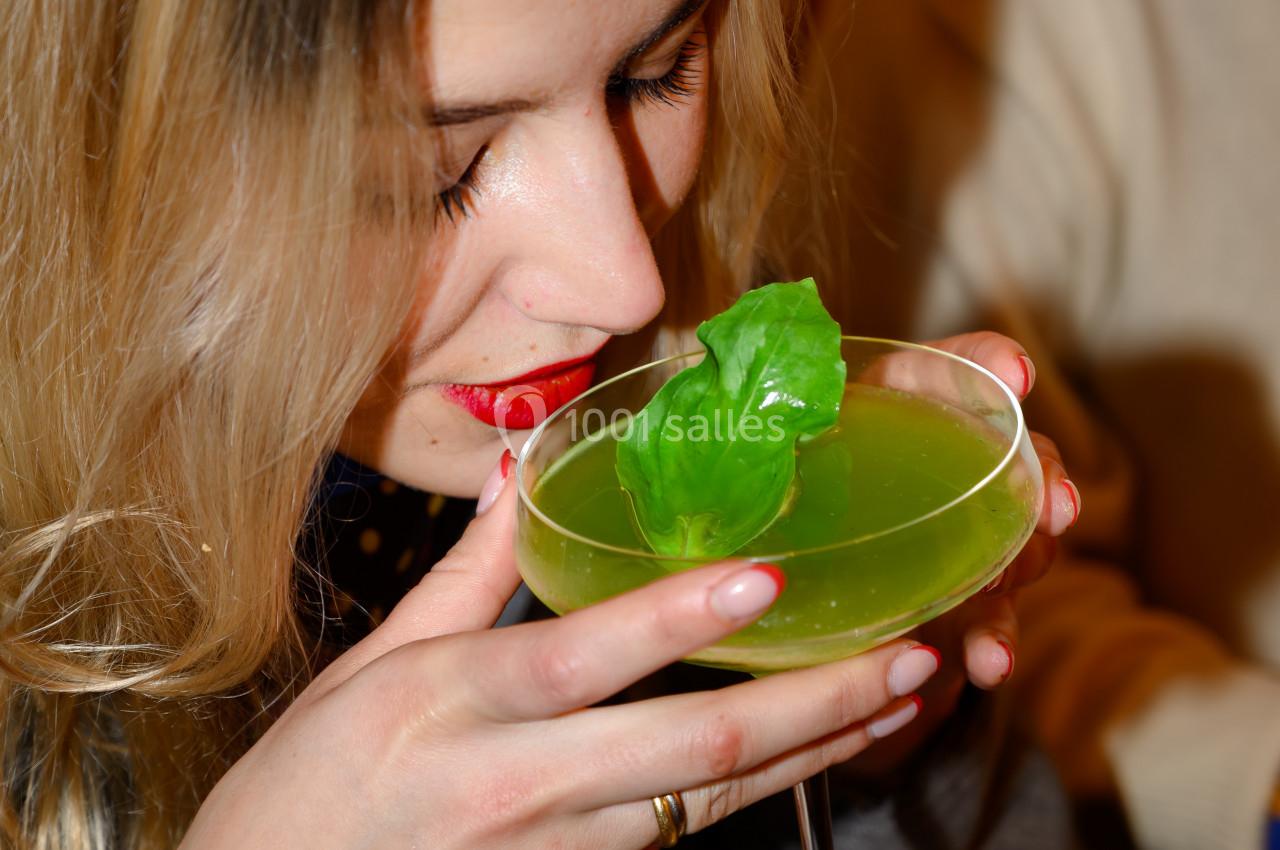 Une femme aux cheveux blonds sent un cocktail vert garni d'une feuille, tenant délicatement le verre.