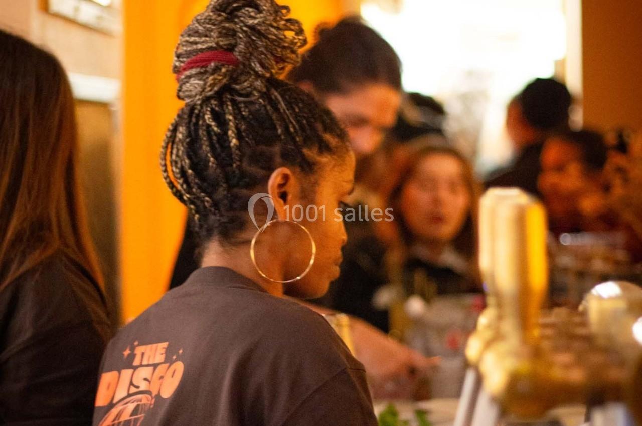Une femme de dos avec des tresses et des boucles d'oreilles se tient dans un espace animé, probablement un bar ou un café.