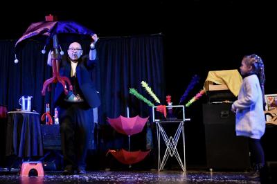 Scène de spectacle avec décorations festives, rideaux noirs, tables couvertes et accessoires colorés sous des éclairages.