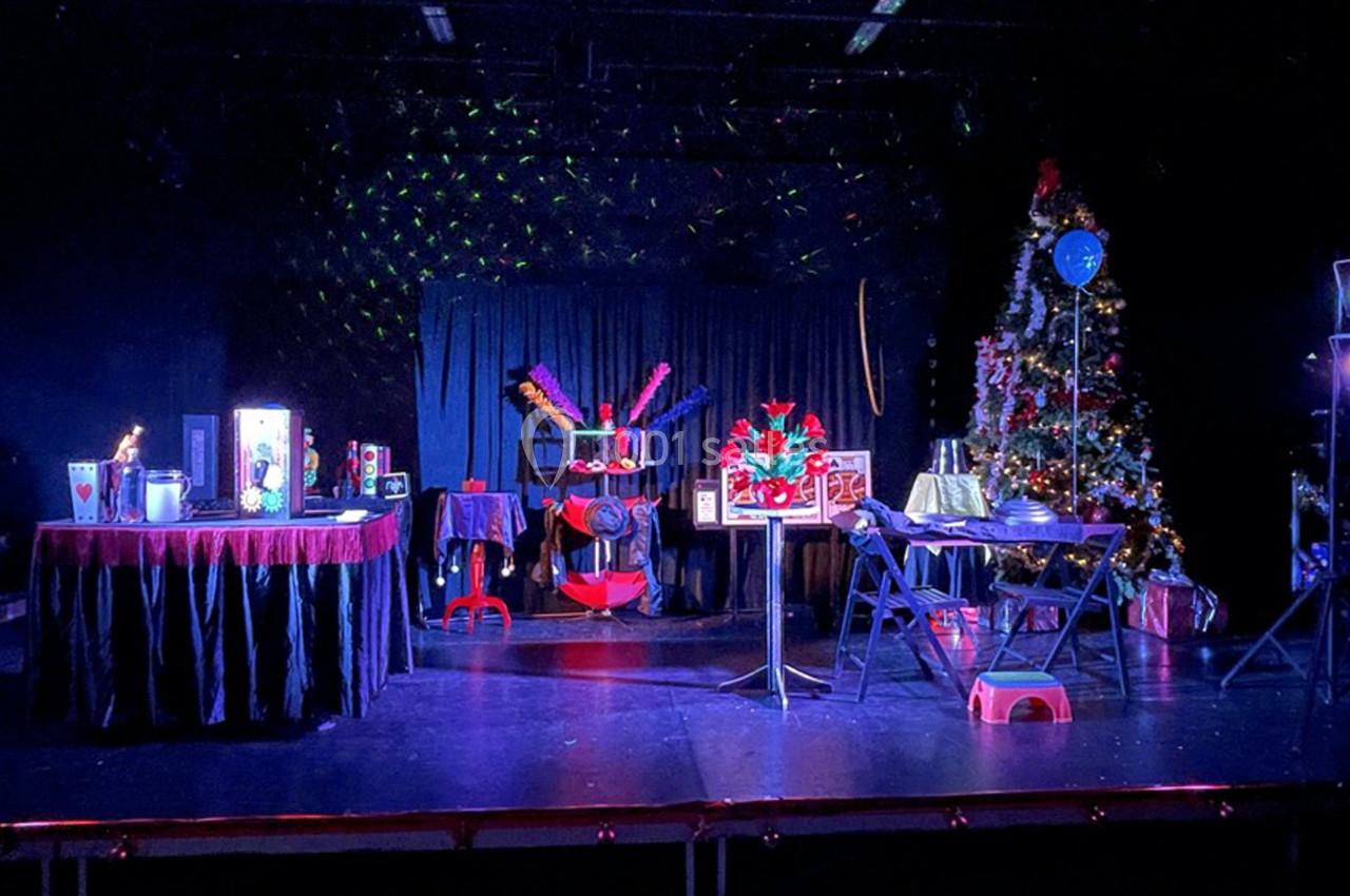 Scène de spectacle avec décorations festives, rideaux noirs, tables couvertes et accessoires colorés sous des éclairages.