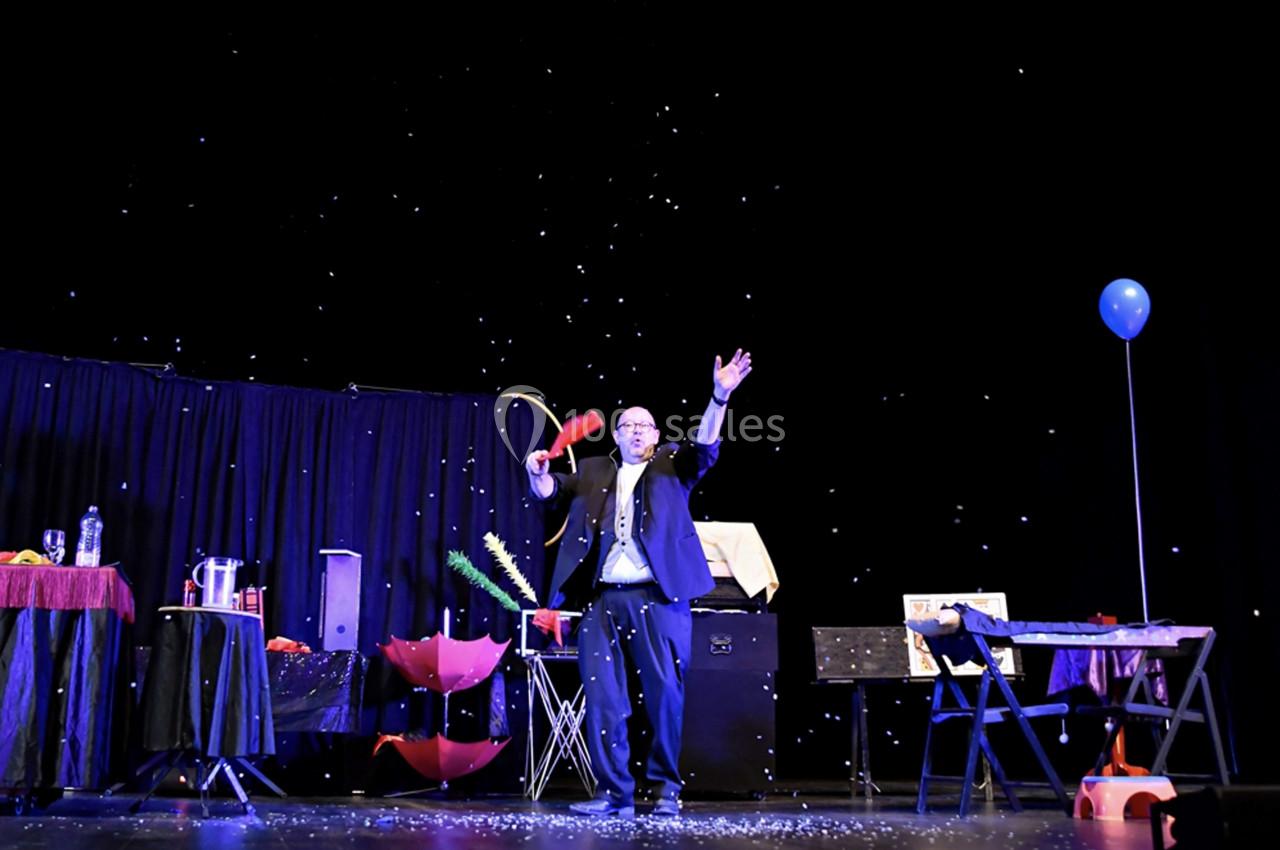 Un magicien en costume réalise un tour sur scène, entouré d'accessoires et de confettis sous un éclairage sombre.