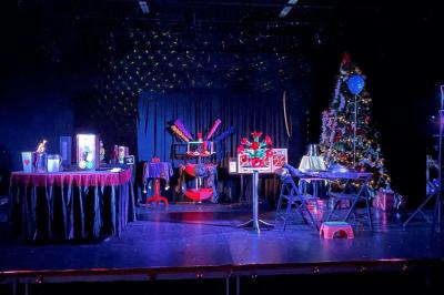 Scène de spectacle avec décorations festives, rideaux noirs, tables ornées et un sapin de Noël illuminé.