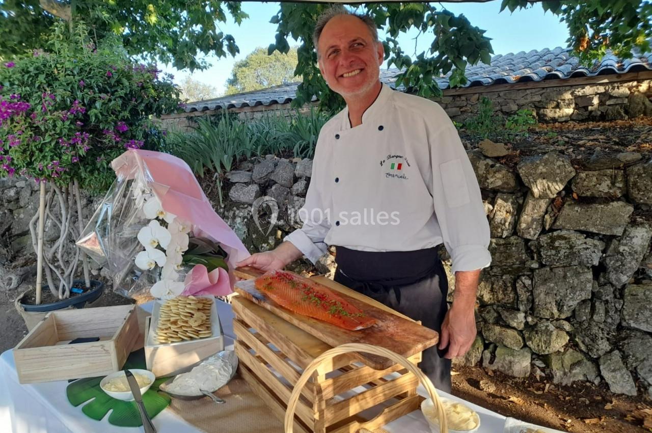 Un chef souriant présente un saumon gravlax sur une table dressée en extérieur, entourée de décorations naturelles.