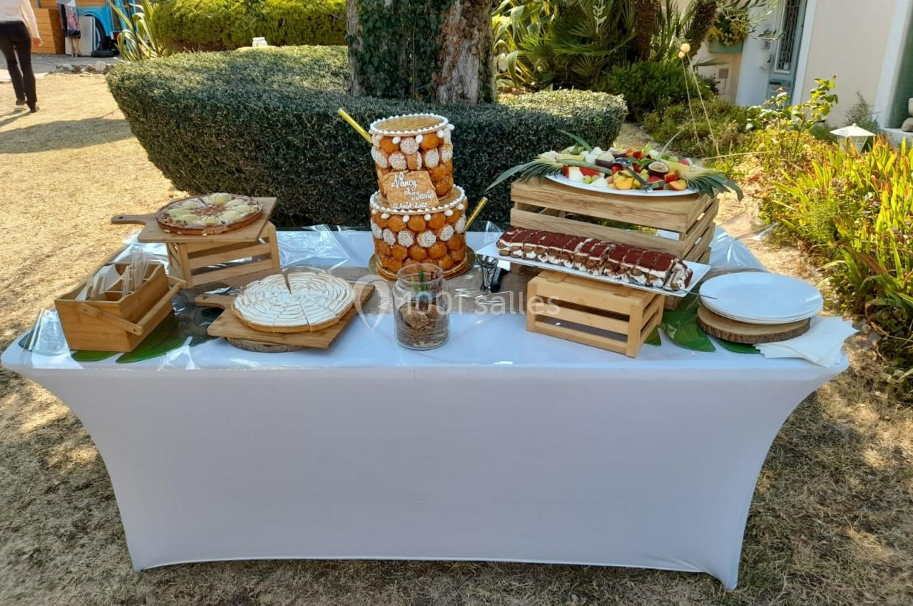 Table de buffet en extérieur avec pâtisseries, fruits frais, croquembouche et plateaux en bois sur une nappe blanche.