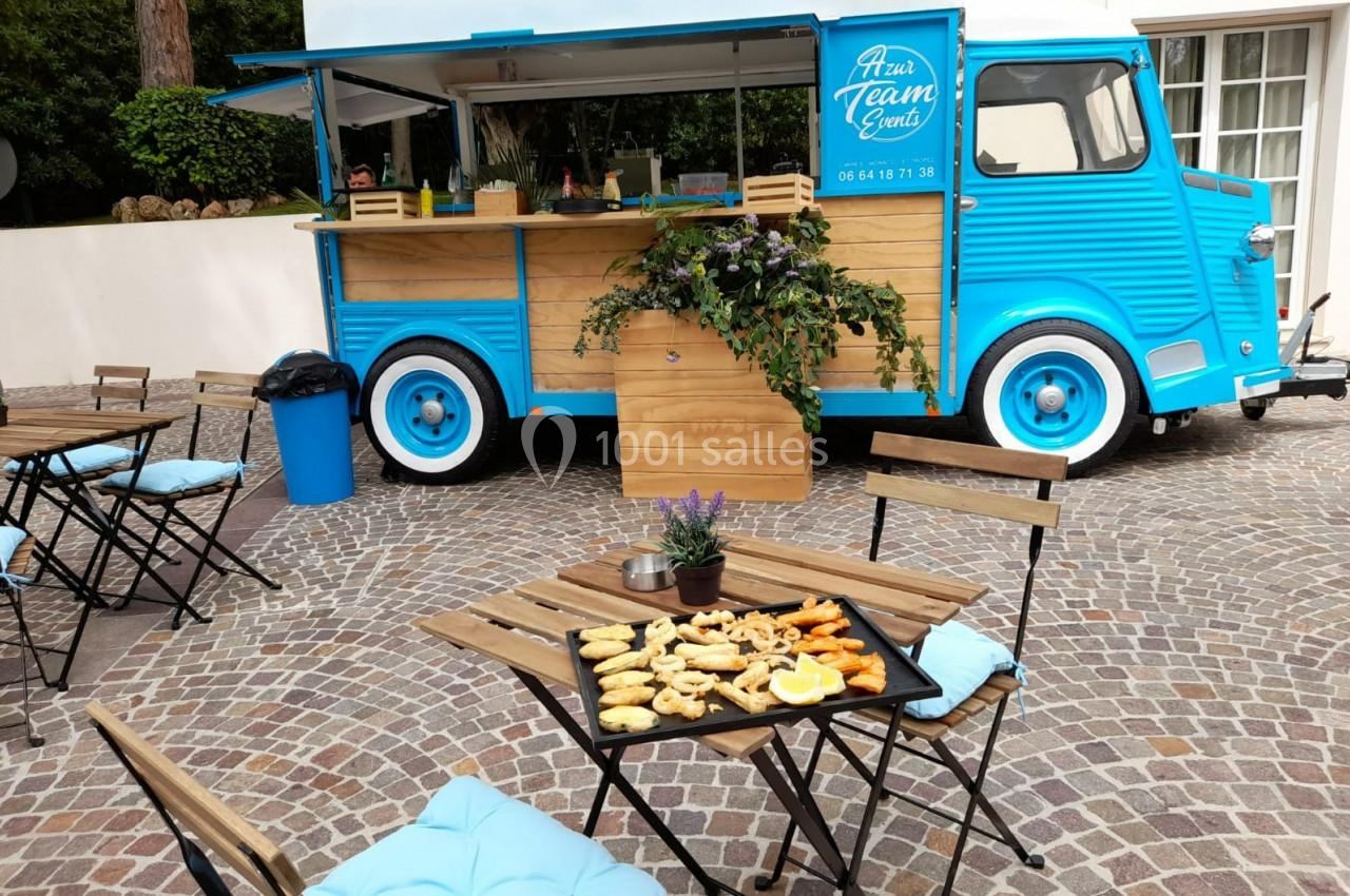 Food truck bleu avec comptoir en bois, tables et chaises en extérieur, plateau de snacks posé sur une table.