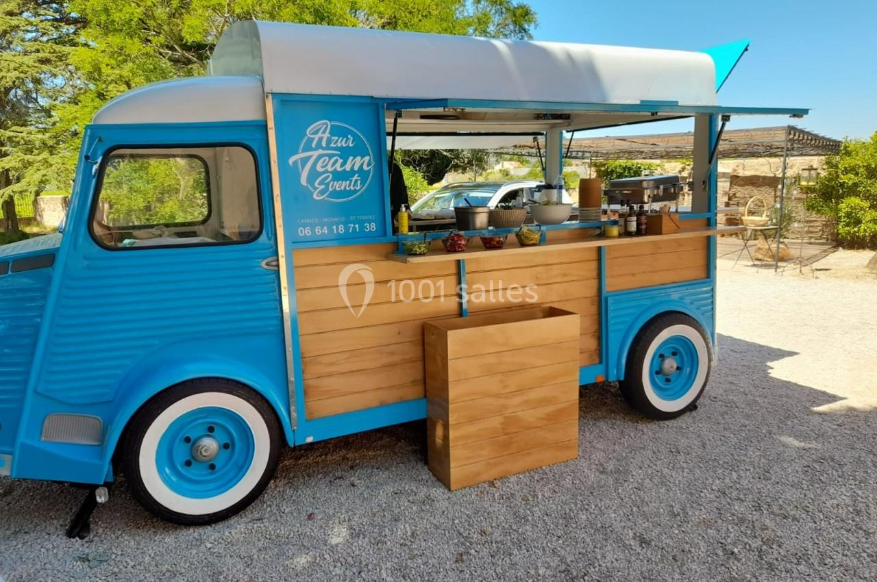 Camion bleu aménagé en food truck avec comptoir en bois, stationné sur un sol gravillonné en extérieur.