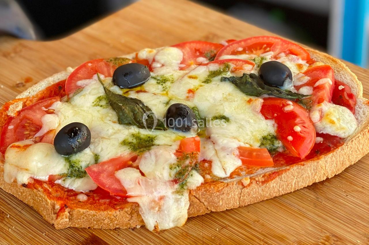 Tranche de pain garnie de tomates, mozzarella fondue, olives noires, basilic et pesto, posée sur une planche en bois.