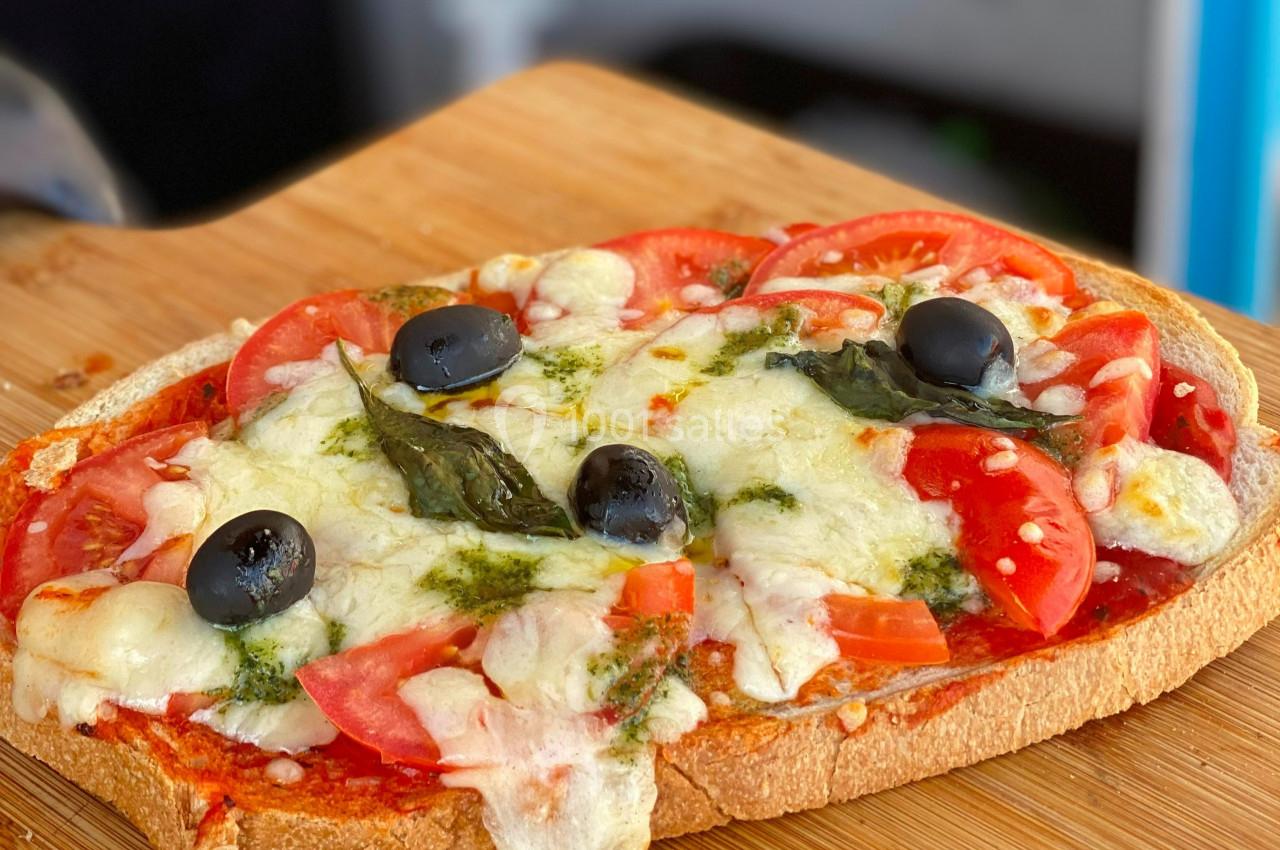 Tranche de pain garnie de tomates, fromage fondu, olives noires, basilic et sauce pesto sur une planche en bois.