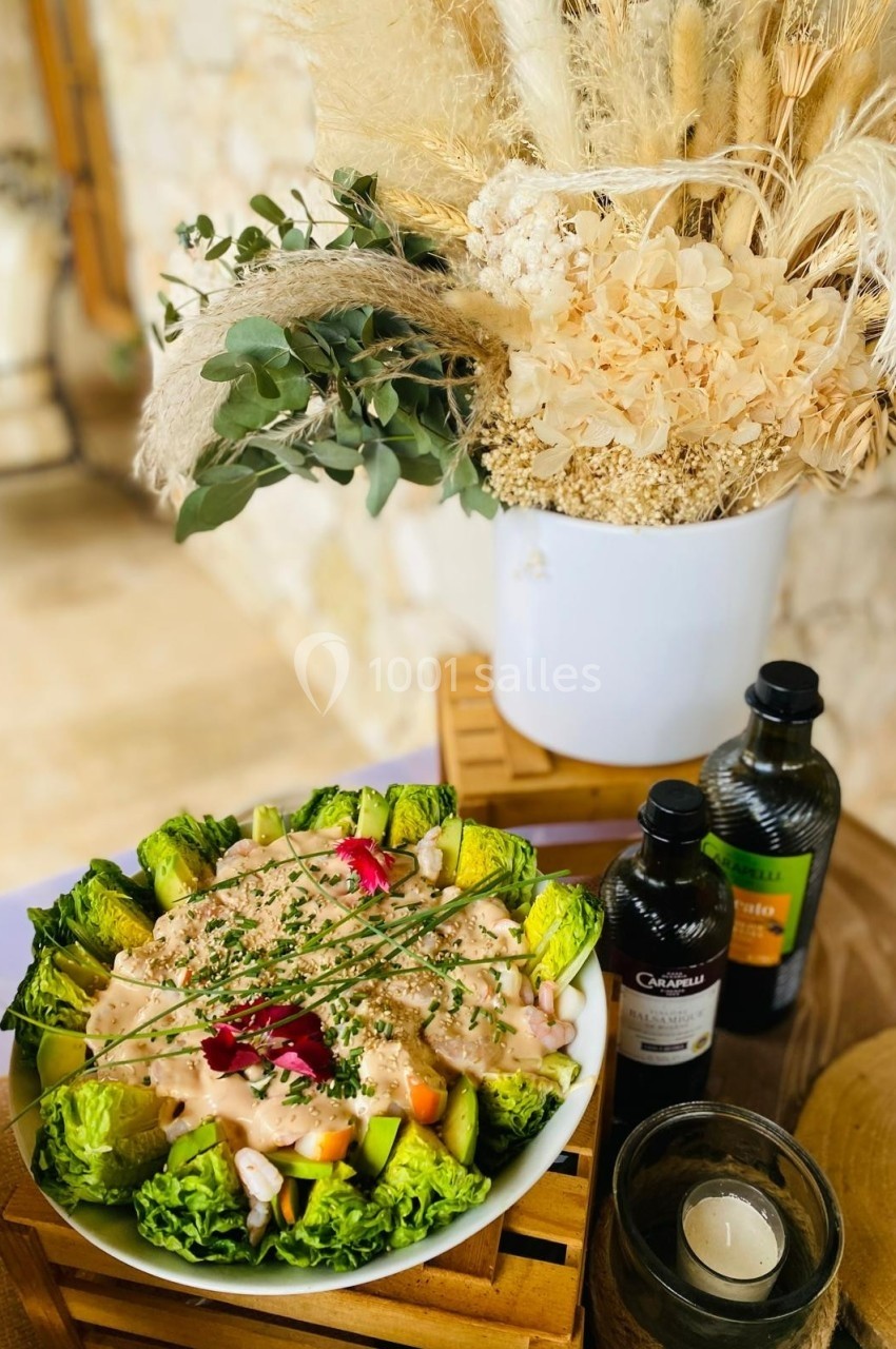 Salade garnie de légumes frais et sauce, présentée sur une table en bois avec décorations végétales et bouteilles d'huile.