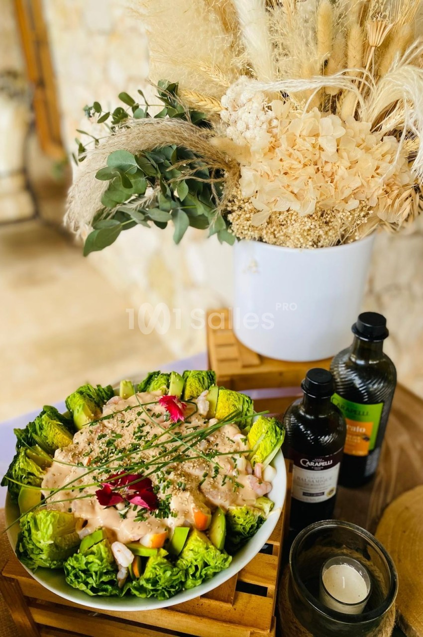 Salade garnie de légumes frais et sauce, présentée sur une table en bois avec décorations végétales et bouteilles d'huile.