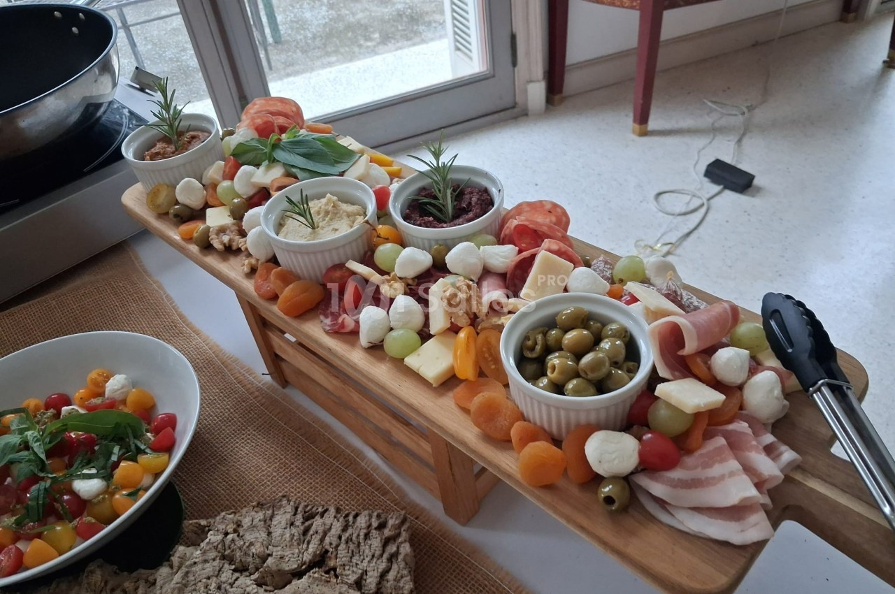 Plateau en bois garni de charcuteries, fromages, olives, fruits secs et légumes, avec des ramequins au centre.