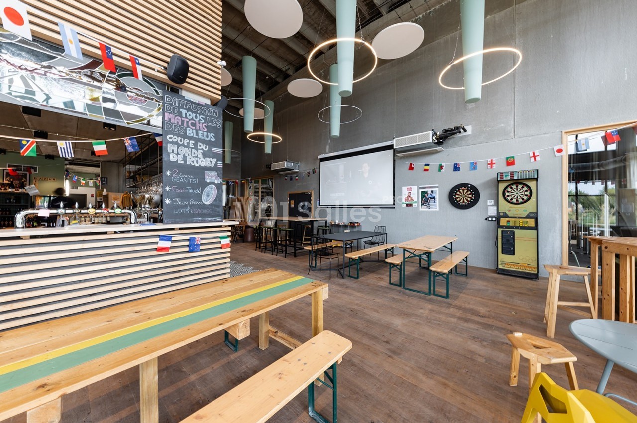 Intérieur d'un bar avec des tables en bois, un écran de projection, un tableau noir et des décorations de drapeaux.