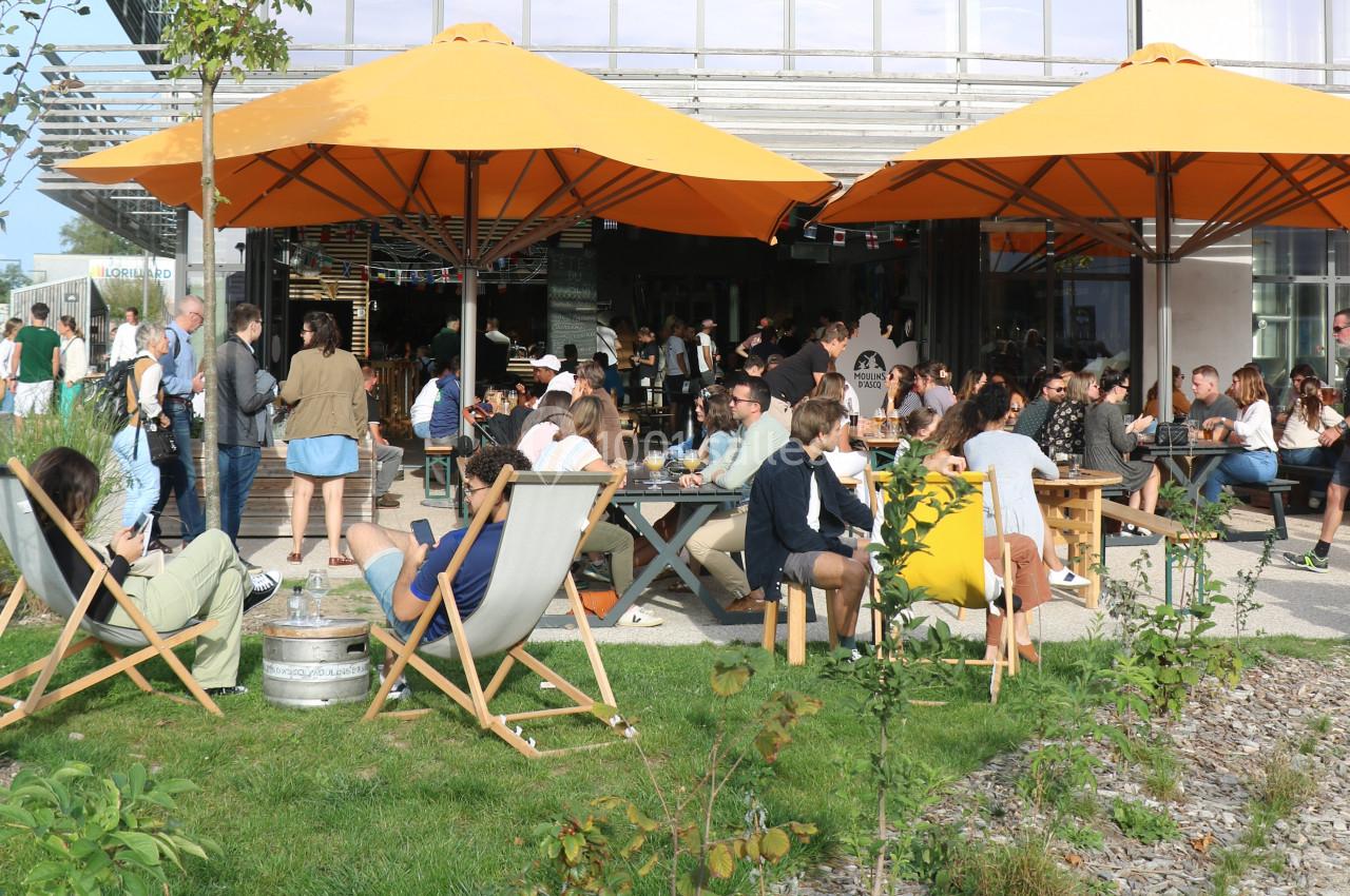 Personnes assises sous des parasols orange dans un espace extérieur, profitant d'un moment convivial par beau temps.