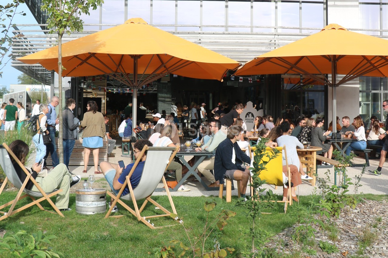 Terrasse animée avec des tables et chaises longues, des personnes profitant du soleil sous des parasols orange.