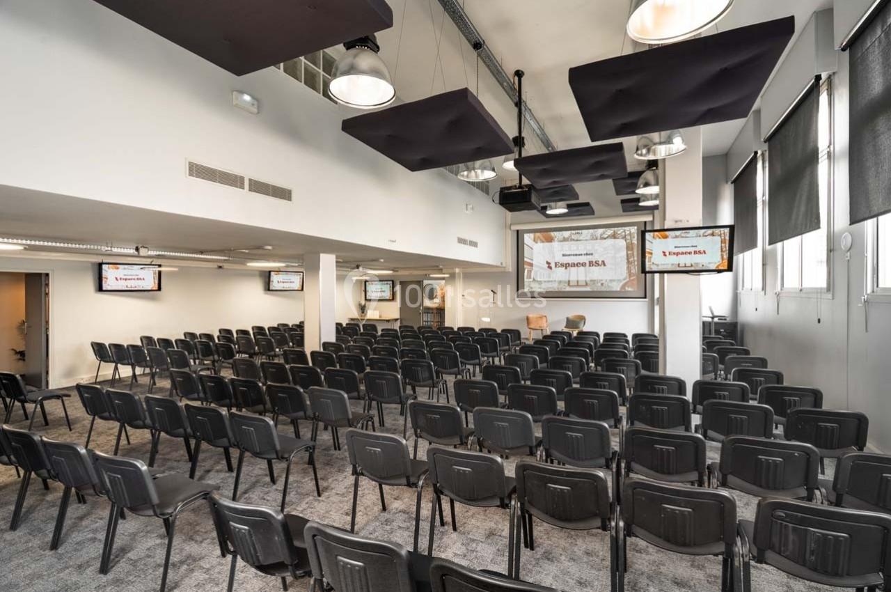 Salle de conférence lumineuse avec rangées de chaises noires, écrans et panneaux acoustiques suspendus au plafond.