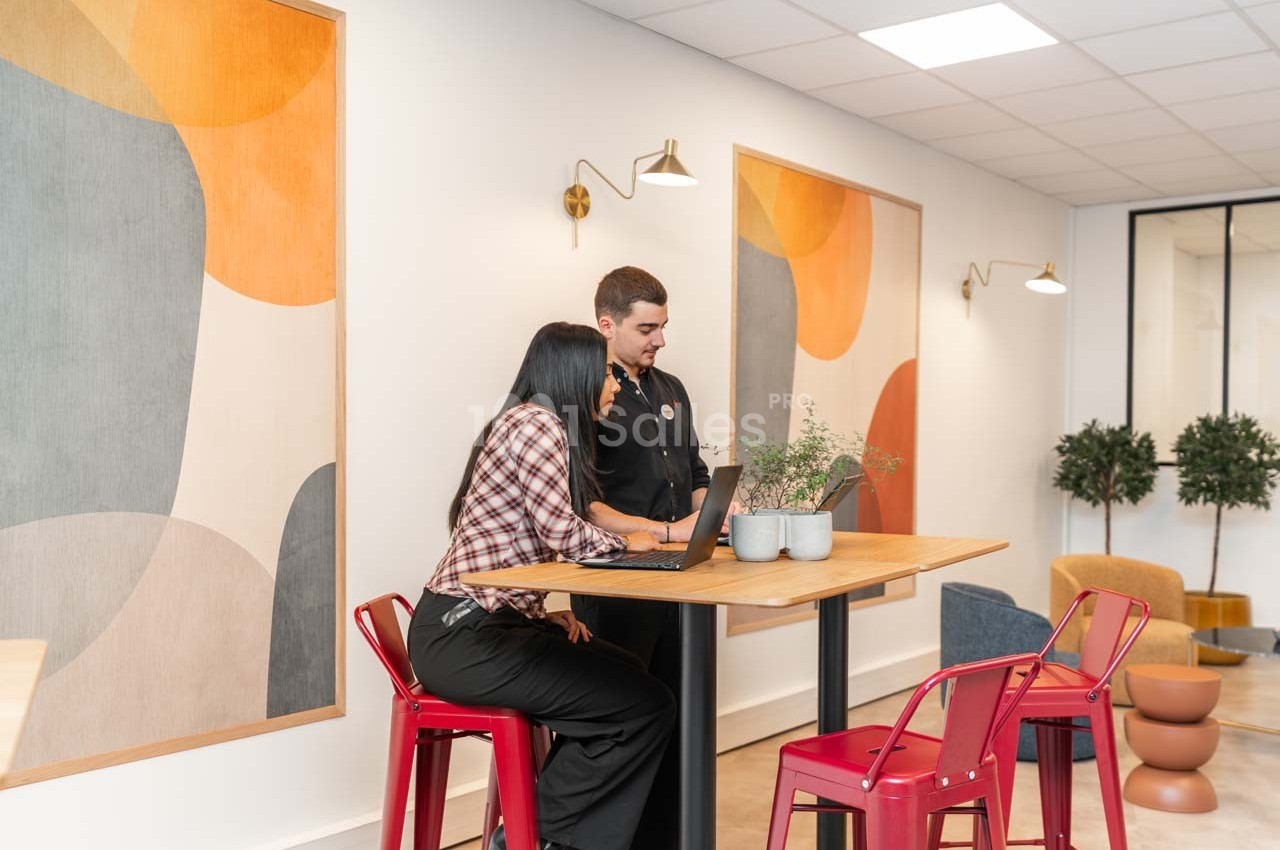 Deux personnes discutent devant un ordinateur portable dans un espace moderne avec des décorations murales colorées.