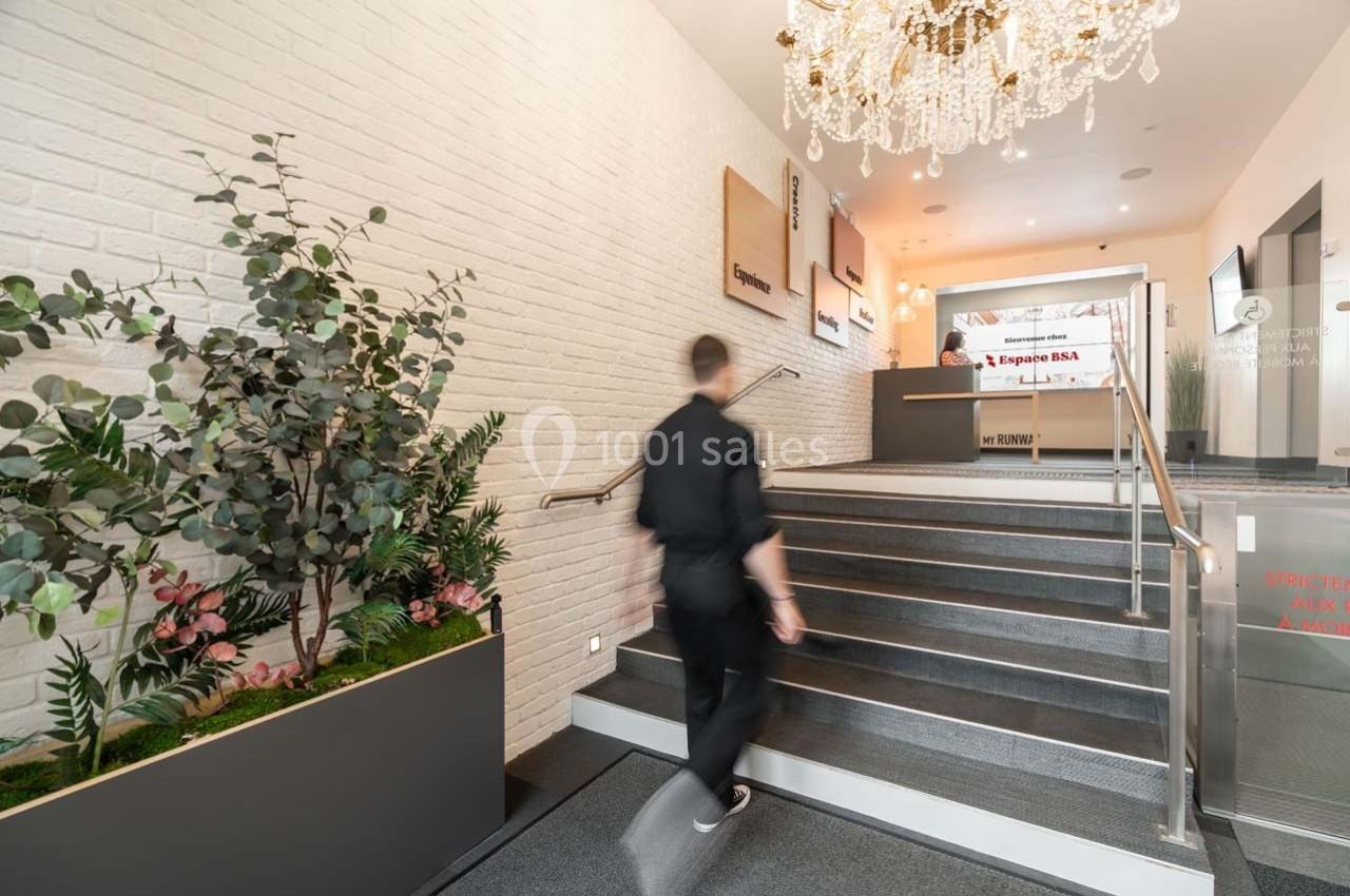Un homme monte un escalier dans un hall lumineux avec des plantes, un mur en briques blanches et un lustre.