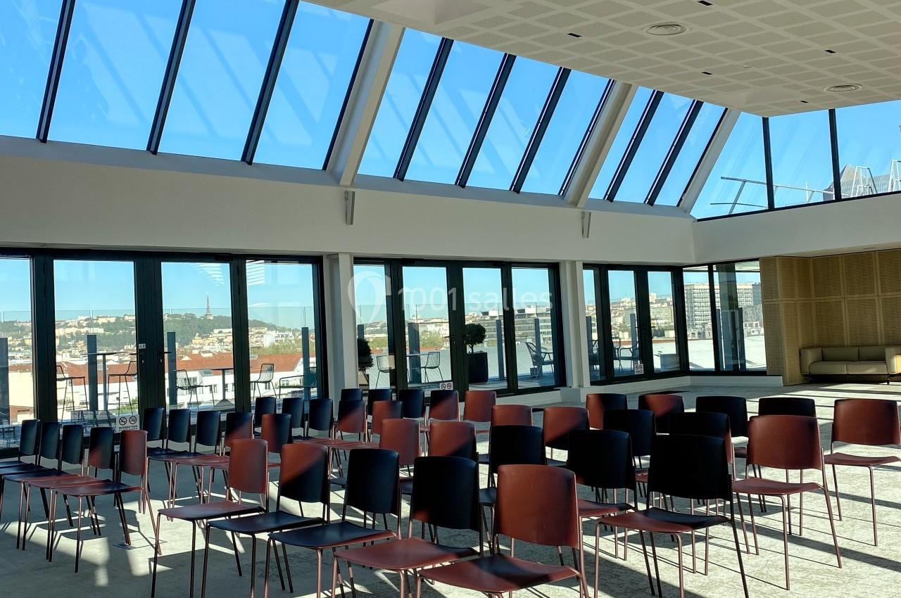 Salle lumineuse avec de grandes baies vitrées, des rangées de chaises alignées et vue sur des bâtiments en arrière-plan.