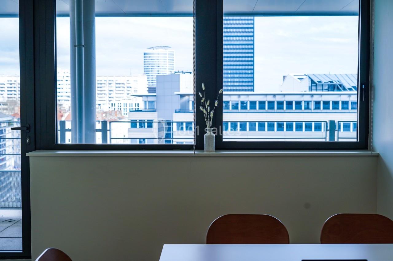 Vue depuis une salle de réunion avec une table, des chaises et une fenêtre donnant sur des immeubles modernes.