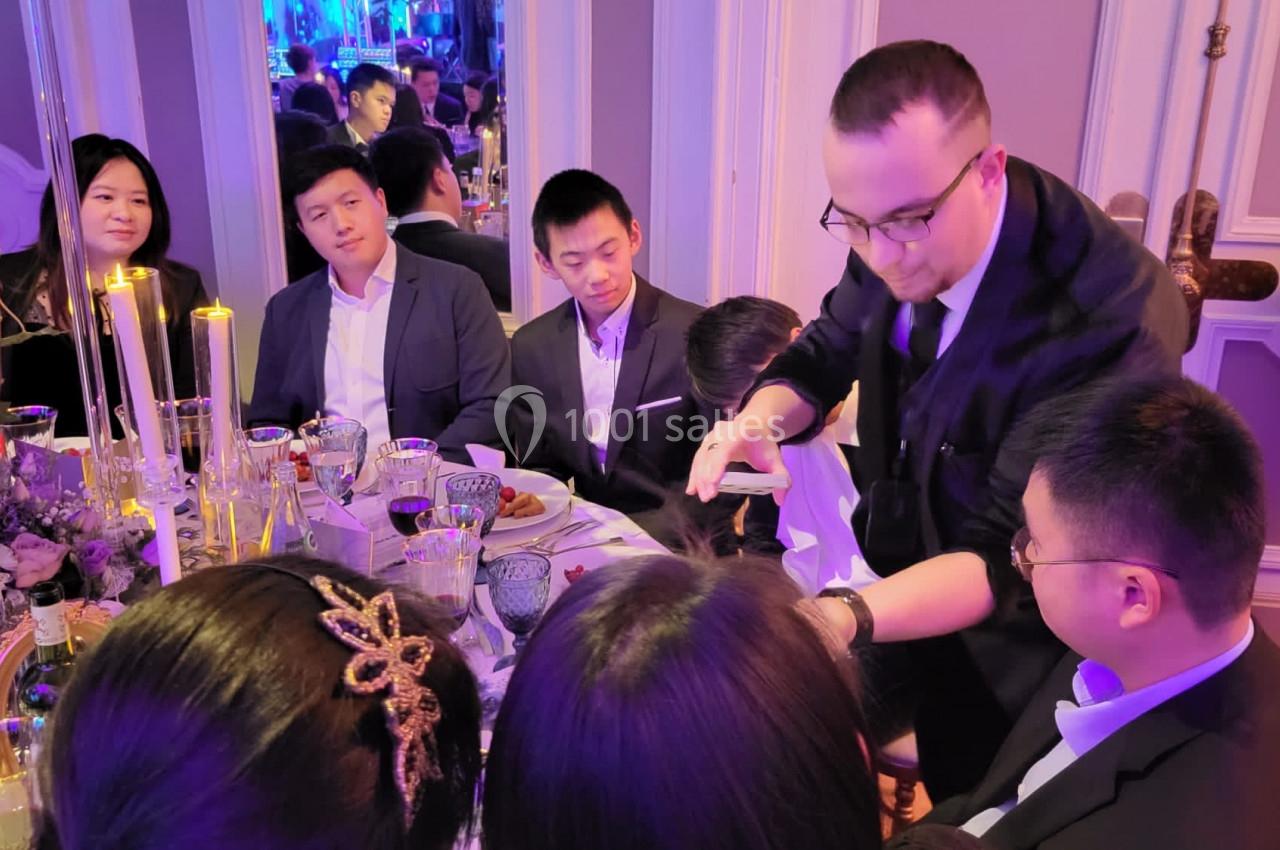 Un magicien réalise un tour de cartes à une table entourée de convives élégamment vêtus lors d'un dîner.