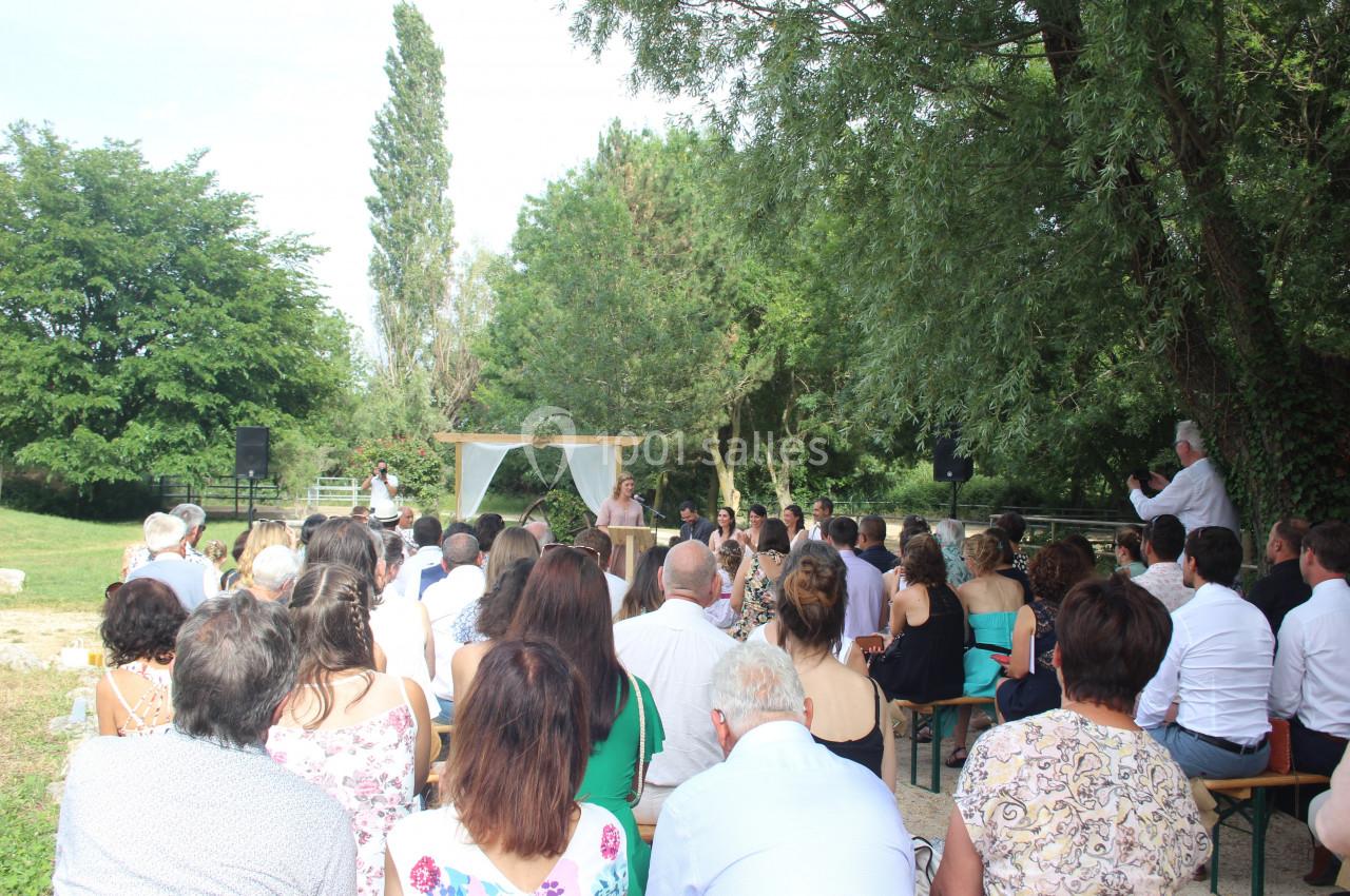 Un groupe de personnes assiste à une cérémonie en plein air, sous des arbres, avec un officiant devant une arche décorée.