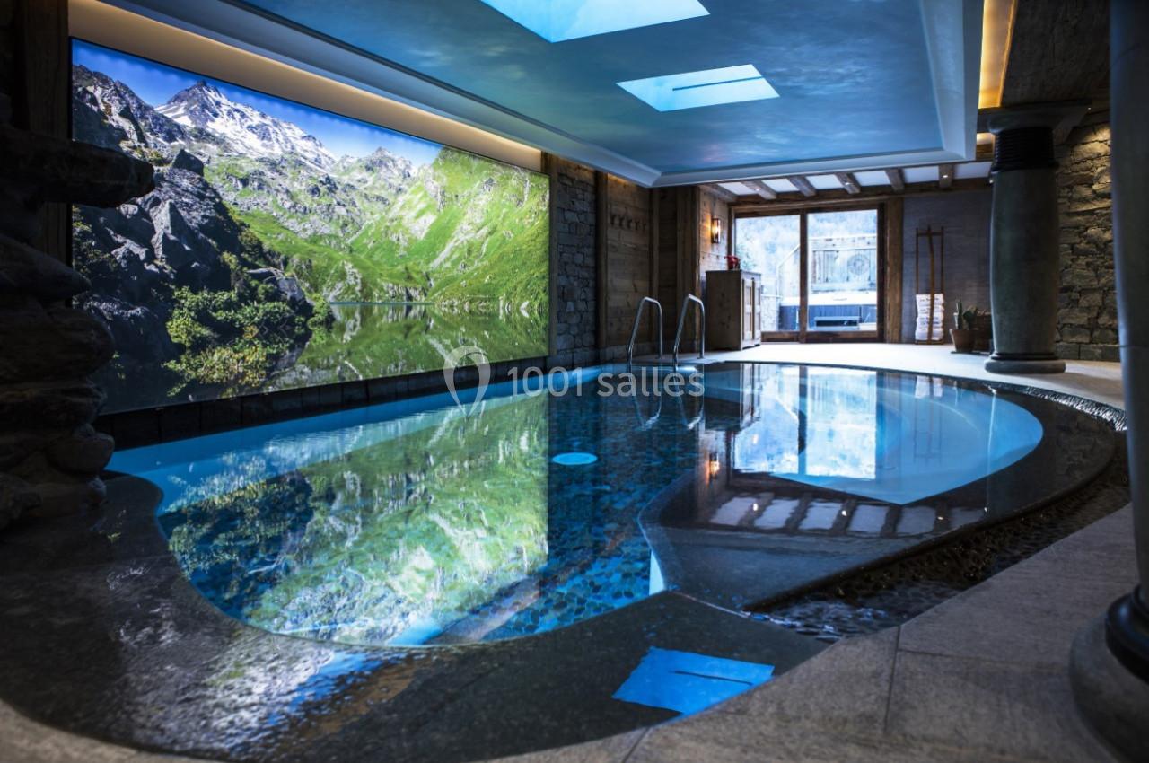 Piscine intérieure avec eau claire, entourée de murs en pierre, éclairée par des puits de lumière et une fresque alpine.