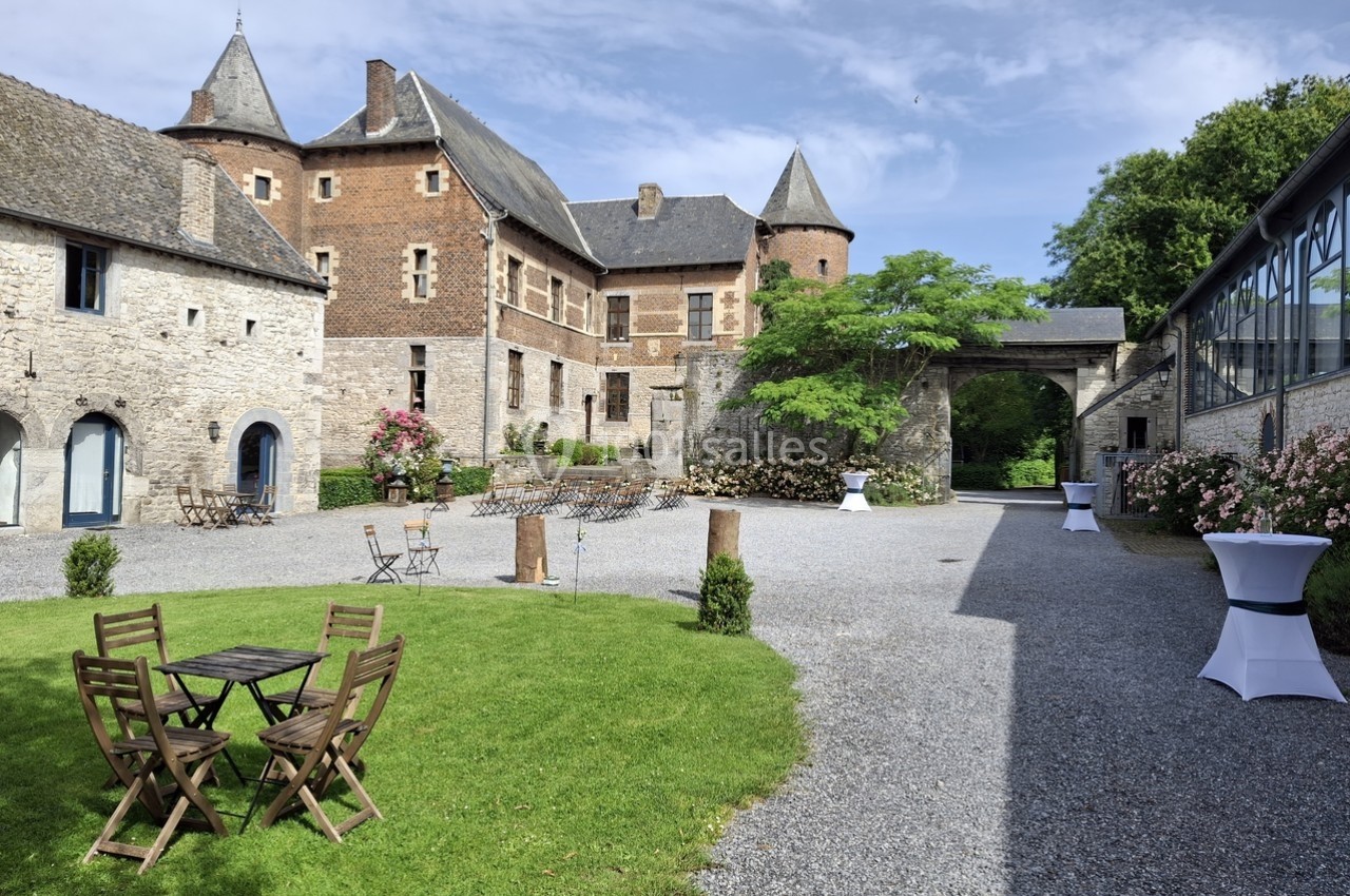 Cour d'un château historique avec pelouse, mobilier de jardin en bois, allées gravillonnées et bâtiments en pierre.