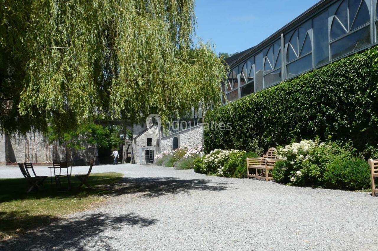 Allée gravillonnée bordée de bancs, de fleurs et de verdure, près d'un bâtiment vitré sous un ciel bleu.